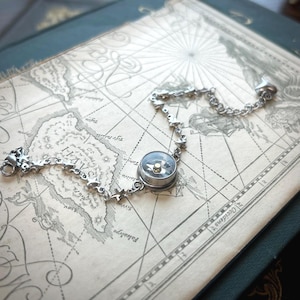 May include: Silver-toned bracelet featuring a compass design. The bracelet includes a small compass with a clear cover and a chain adorned with star-shaped charms. The bracelet is displayed on a vintage map.