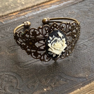 May include: A delicate bronze bracelet with an ornate filigree design. A black cameo with a white rose is set in the center of the bracelet.