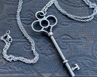 May include: A silver-colored key pendant necklace. The key has an ornate top with a long stem and a T-shaped end. The chain is a delicate, linked design. The necklace is displayed on a textured, dark surface.