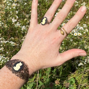 May include: A bronze-toned cuff bracelet featuring an oval black cameo with a cream-colored cat silhouette, a matching oval cameo ring, and a bronze-toned snake-shaped ring with a textured scale pattern on the band.