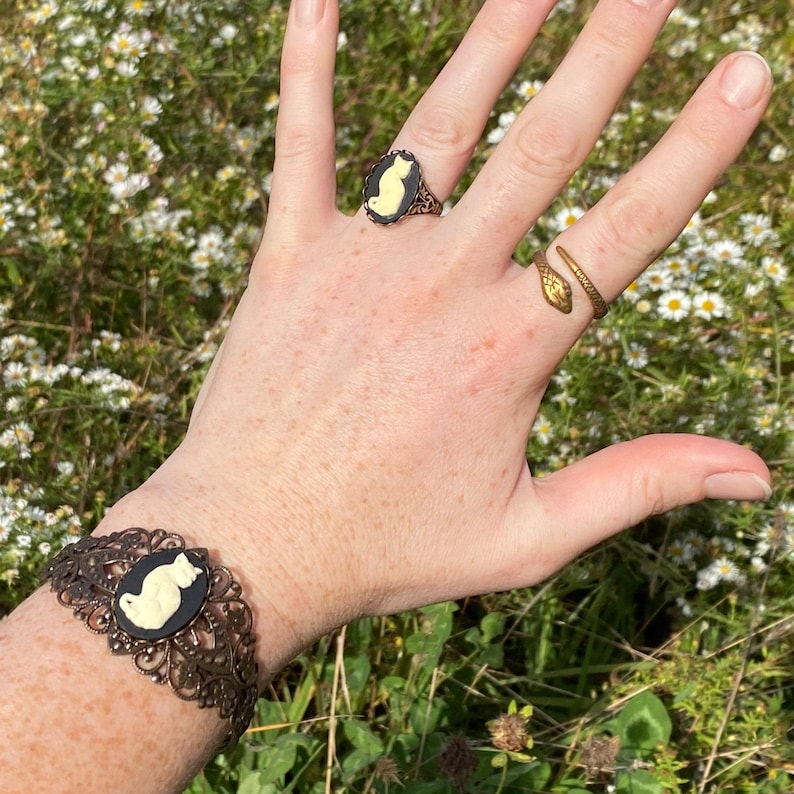 May include: A bronze-toned cuff bracelet featuring an oval black cameo with a cream-colored cat silhouette, a matching oval cameo ring, and a bronze-toned snake-shaped ring with a textured scale pattern on the band.