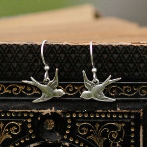 May include: Silver dangle earrings featuring a pair of stylized silver birds with wings outstretched. The earrings are hanging from a black background.