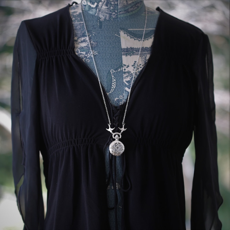 May include: A silver necklace with a pocket watch pendant and two bird charms. The necklace is on a black sheer top.