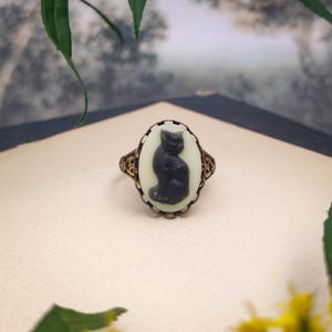 May include: An oval ring featuring a black cat silhouette cameo set against a cream-colored background. The ring has an ornate, antiqued bronze-toned metal setting with intricate filigree detailing along the band and the oval frame.