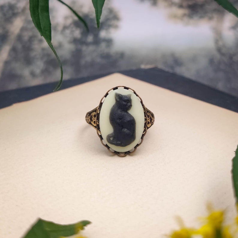 May include: An oval ring featuring a black cat silhouette cameo set against a cream-colored background. The ring has an ornate, antiqued bronze-toned metal setting with intricate filigree detailing along the band and the oval frame.