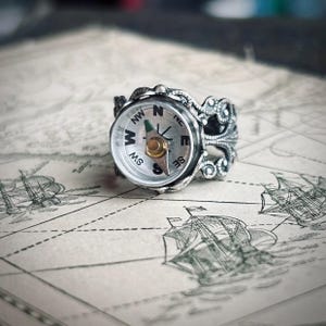 May include: A silver compass ring with an ornate design. The ring features a working compass with a white face, black lettering, and a gold needle. The ring is set on a map background with illustrations of sailing ships.