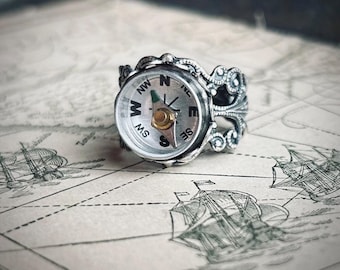May include: A silver compass ring with an ornate design. The ring features a working compass with a white face, black lettering, and a gold needle. The ring is set on a map background with illustrations of sailing ships.