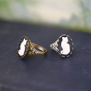 May include: Two rings featuring oval cameos with white cat silhouettes on black backgrounds. One ring has a brass-toned metal band with intricate filigree detailing, while the other has a silver-toned metal band with similar ornate patterns.