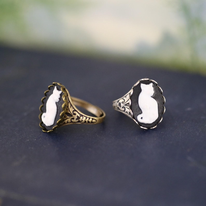 May include: Two rings featuring oval cameos with white cat silhouettes on black backgrounds. One ring has a brass-toned metal band with intricate filigree detailing, while the other has a silver-toned metal band with similar ornate patterns.