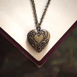 Puede incluir: Un colgante de relicario en forma de corazón hecho de metal con tono de latón antiguo, con un patrón floral y de volutas en relieve con un corazón liso en el centro. El colgante cuelga de un collar de cadena de eslabones a juego.