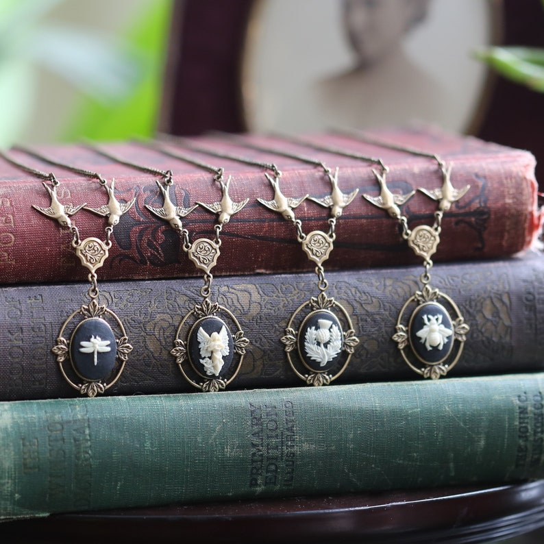 May include: Five delicate necklaces with gold chains and pendants featuring black cameo-like settings with white cameo designs of a dragonfly, angel, thistle, and bee. Each necklace has a pair of gold birds on the chain.