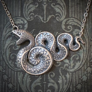 May include: A silver-toned snake pendant necklace. The snake is coiled with an open mouth, and detailed scales. The necklace chain is also silver. The pendant is set against a dark, patterned background.