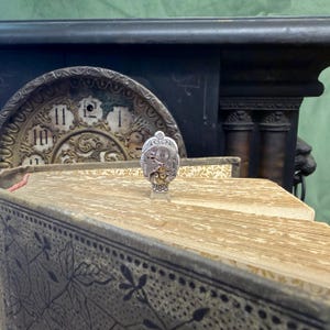 Puede incluir: Un peque&ntilde;o mecanismo de reloj plateado, con engranajes intrincados, se muestra sobre un libro antiguo. El libro tiene p&aacute;ginas envejecidas y una cubierta decorativa con patrones florales. El fondo incluye una esfera de reloj vintage.
