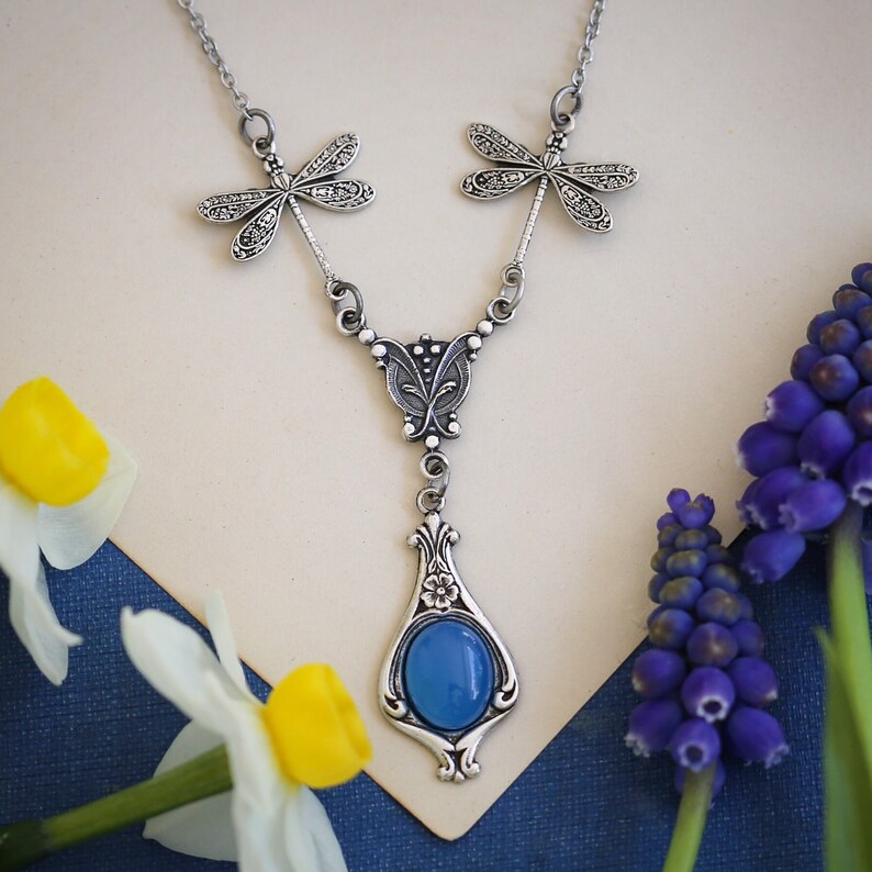 May include: A silver necklace with a blue gemstone pendant and dragonfly charms. The necklace is on a white background with blue flowers.