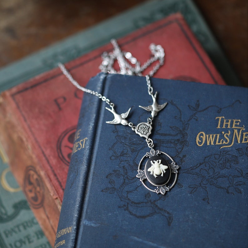 May include: A silver necklace with a black cameo featuring a white bee. The necklace has two silver birds flying upwards and a floral design. The necklace is displayed on top of two vintage books.