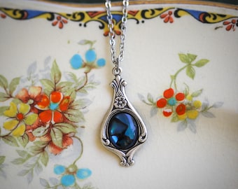 Pode incluir: Um colar de prata com um pingente em forma de lágrima que apresenta uma pedra azul escura iridescente. O pingente tem um design floral e está colocado sobre um fundo branco com padrões florais.