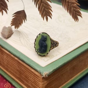 May include: A ring featuring an oval cameo with a dark blue cat silhouette set against a light green background. The metal band is finished in an aged bronze tone with intricate filigree detailing along the sides of the setting.