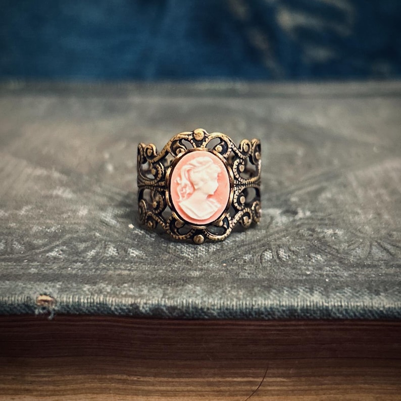 Pode incluir: Um anel de ouro com um delicado design de filigrana e um camafeu com o perfil de uma mulher em rosa.