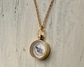 May include: A gold compass necklace with a small round compass pendant. The compass face has black markings and a blue needle.