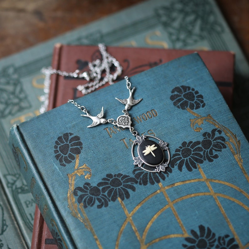 May include: A silver necklace with a dragonfly pendant. The pendant is black with a white dragonfly and is set in a silver frame. The necklace is on top of a stack of old books with a blue cover.