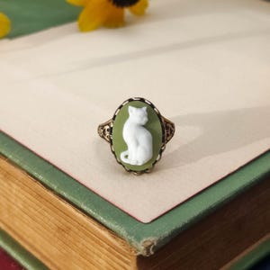 May include: An oval ring featuring a white cat cameo set against a sage green background. The ring has an intricate, dark metal filigree band with decorative scrollwork patterns surrounding the central oval setting.