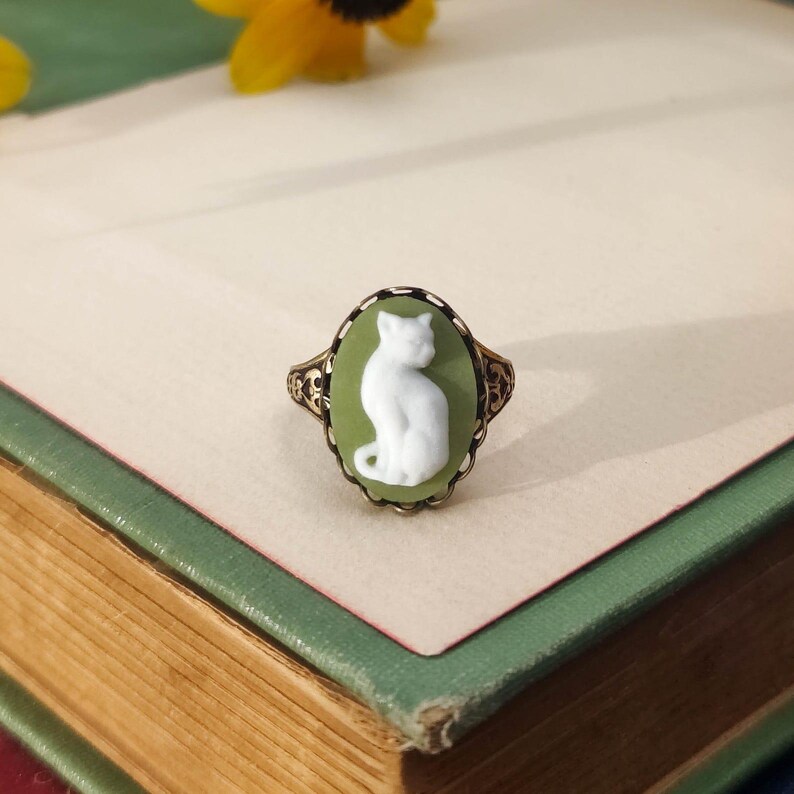 May include: An oval ring featuring a white cat cameo set against a sage green background. The ring has an intricate, dark metal filigree band with decorative scrollwork patterns surrounding the central oval setting.