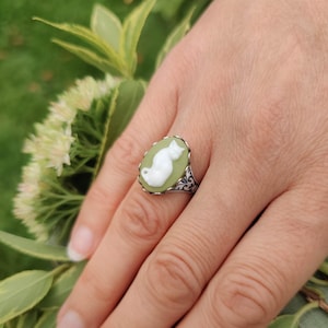 May include: A silver ring featuring an oval-shaped cameo with a light green background and a white relief of a sitting cat. The metal band displays intricate scrollwork patterns along the sides, framing the central cat motif clearly.