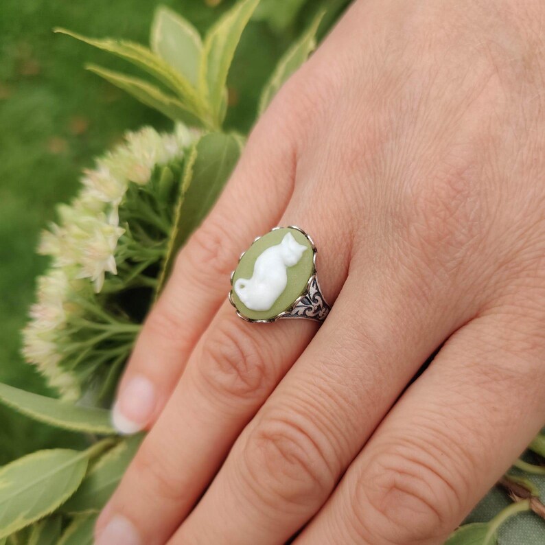 May include: A silver ring featuring an oval-shaped cameo with a light green background and a white relief of a sitting cat. The metal band displays intricate scrollwork patterns along the sides, framing the central cat motif clearly.