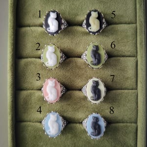 May include: Eight oval rings featuring cat cameos in various color combinations. Ring 1: white cat on black. 2: white on light green. 3: white on pink. 4: white on light blue. 5: cream on black. 6: dark gray on light green. 7: dark gray on cream. 8: dark gray on light blue.