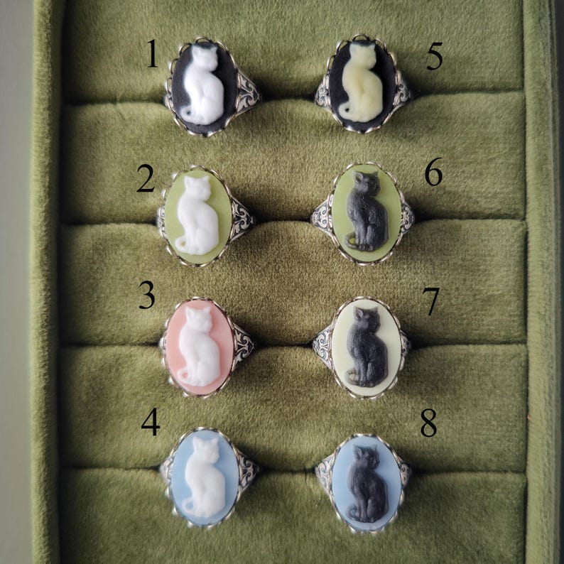 May include: Eight oval rings featuring cat cameos in various color combinations. Ring 1: white cat on black. 2: white on light green. 3: white on pink. 4: white on light blue. 5: cream on black. 6: dark gray on light green. 7: dark gray on cream. 8: dark gray on light blue.