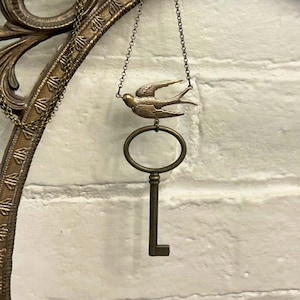 May include: A gold necklace featuring a key charm and a small bird pendant. The necklace is hanging from an ornate metal frame.