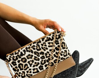 May include: A rectangular clutch bag featuring a black and tan leopard print pattern on the upper section and a solid tan textured base. The bag includes a gold-tone metal chain strap and a black tassel attached to the side zipper pull.