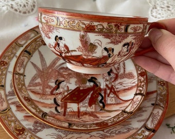 May include: A vintage Japanese teacup and saucer set with a delicate floral and figure design in shades of red, gold, and black. The set features a traditional Japanese aesthetic.
