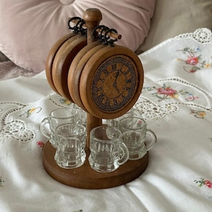 Puede incluir: Soporte de vasos de chupito de madera con una esfera de reloj en la parte superior y seis pequeños vasos de chupito de vidrio. El soporte es marrón y los vasos de chupito son transparentes.