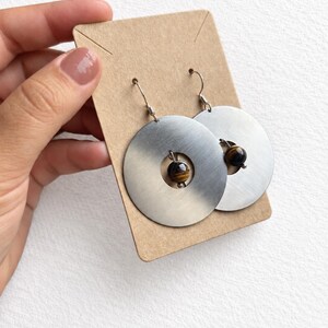 May include: A pair of drop earrings featuring large, circular, brushed silver-tone metal discs. Each disc has a central cutout holding a single, round, brown and gold tiger's eye gemstone bead. The earrings are attached to silver-tone metal hooks.