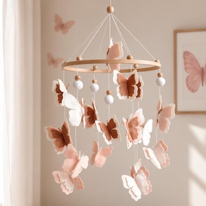 May include: A hanging nursery mobile featuring multiple felt butterflies in shades of dusty rose, terracotta, and cream. The butterflies are suspended from a circular wooden hoop by thin white strings, interspersed with small round wooden and white felt beads.