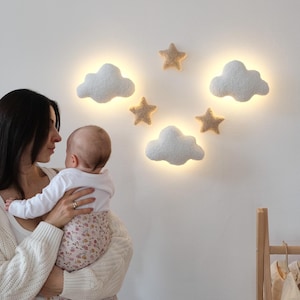 Peut inclure: Décoration murale de chambre d'enfant avec trois lampes en forme de nuages blancs et quatre étoiles beiges. Les lumières sont disposées sur un mur blanc, créant une lueur douce et chaleureuse. Thème : chambre d'enfant fantaisiste et rêveuse.
