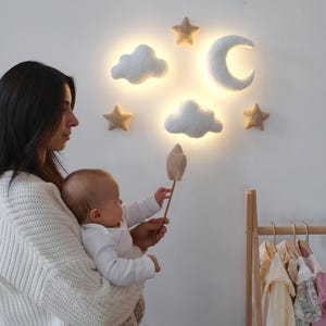 Peut inclure: Une applique murale de chambre d'enfant avec une lune en croissant, des étoiles et des nuages. La lumière est placée sur un mur blanc. Le design imite un ciel nocturne, offrant une douce lumière ambiante.
