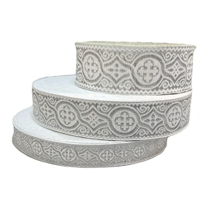 Puede incluir: Tres rollos apilados de cinta decorativa. La cinta tiene una base blanca con un patrón gris adornado con cruces y diseños en espiral. Los rollos son de diferentes tamaños, creando un efecto escalonado.