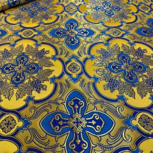 May include: Close-up of a decorative fabric with a repeating pattern. The fabric features a yellow background with gold and blue floral and cross designs. The pattern includes stylized grape clusters and leaf motifs.