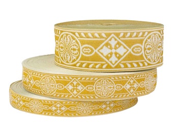 May include: Three spools of decorative ribbon. The ribbon has a gold background with a repeating white geometric pattern. The pattern includes floral and diamond shapes. The spools are stacked on top of each other.