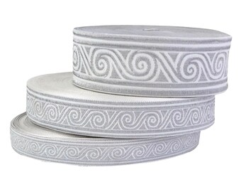 May include: Three rolls of decorative ribbon in varying sizes. The ribbon is white with a silver, repeating scroll pattern. The rolls are stacked on top of each other, with the largest roll on top.