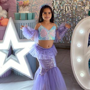 May include: A young person in a mermaid costume poses at a birthday party. The costume includes a sequined top and skirt, with sheer purple sleeves and a tulle skirt. A large illuminated number six and star-shaped light are in the background, along with balloons.