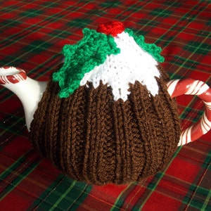May include: A ceramic teapot with a brown knitted cozy shaped like a Christmas pudding. The cozy has white icing, green holly leaves, and a red berry on top. The handle is a red and white candy cane design.