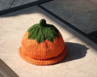 May include: A knitted orange hat with a green leaf-like design on top. The hat has a brim and is made of a textured yarn. The hat is designed to look like a pumpkin.
