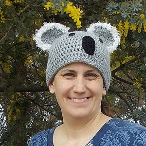 May include: A gray crocheted koala bear hat with white ear accents and a black nose. The hat is worn by a person in front of a background of yellow flowers and green leaves. The hat is handmade.