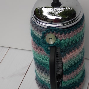 May include: A colorful, handmade crochet coffee pot cozy. The cozy features a button closure and is made with shades of teal, pink, gray, and white yarn. The cozy is designed to fit a coffee pot and keep the contents warm.