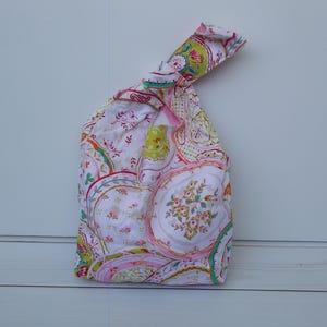 May include: A fabric knot bag with a colourful paisley pattern. The bag features a white background with pink, green, and yellow floral and paisley designs. The bag is knotted at the top for closure.