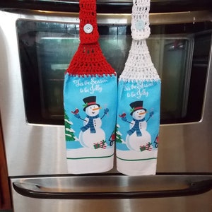 May include: Two kitchen hand towels with a snowman design. The towels have a blue background with a snowman wearing a top hat and scarf. The towels are attached to a red and white crocheted loop with a button.