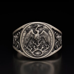 May include: A silver ring with a detailed design of a bird rising from a tree with flames. The ring has a textured surface and intricate details.
